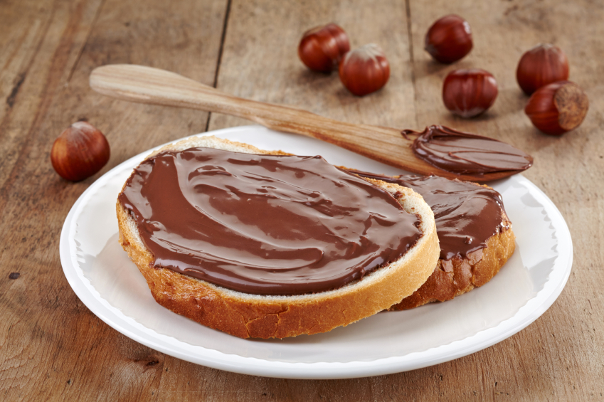 Crema spalmabile al cacao e nocciola su due fette di pane poggiate su un piattino, con un cucchiaio di crema e nocciole sullo sfondo; concept: junk food, zucchero