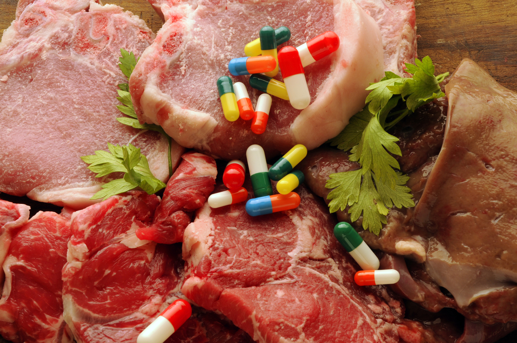 Capsule di farmaci o integratori sparse sopra fette di carne bovina e suina; concept: antibiotici, antibiotico-restistenza