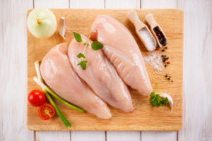 Petti di pollo crudi su un tagliere di legno con cipolla, aglio, cipollotto, erbe, pomodorini, sale e pepe