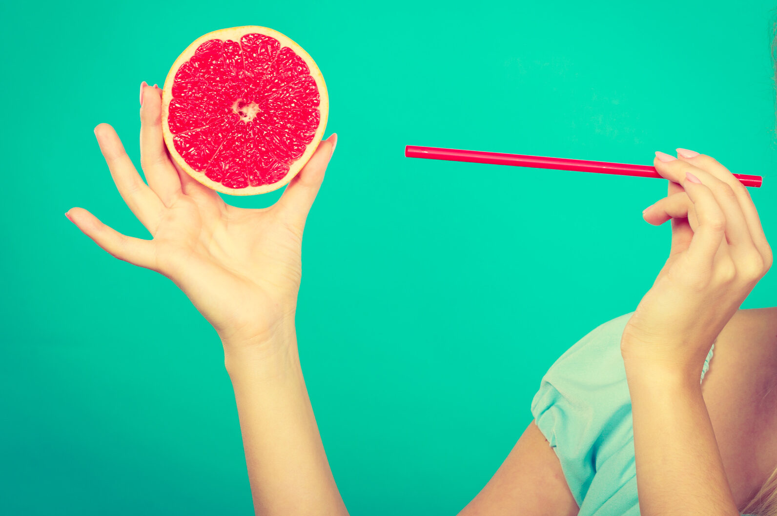 Una donna tiene con una mano mezzo pompelmo e con l'altra una cannuccia; concept: succhi di frutta, centrifughe, estratti