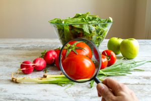 Persona osserva con lente di ingrandimento pomodori su un tavolo insieme a ravanelli, insalata, mele e cipollotti; concept pesticidi