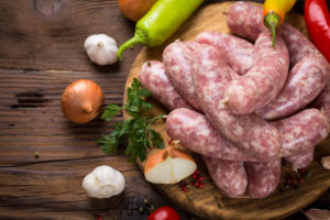 Salsicce crude su un piatto di legno rotondo circondate da aglio, cipolle, peperoncini e prezzemolo