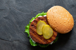 Freshly grilled plant based burger patty in bun with lettuce, slices of gherkin and sauce isolated on black slate. Top view. Copy space. fake meat