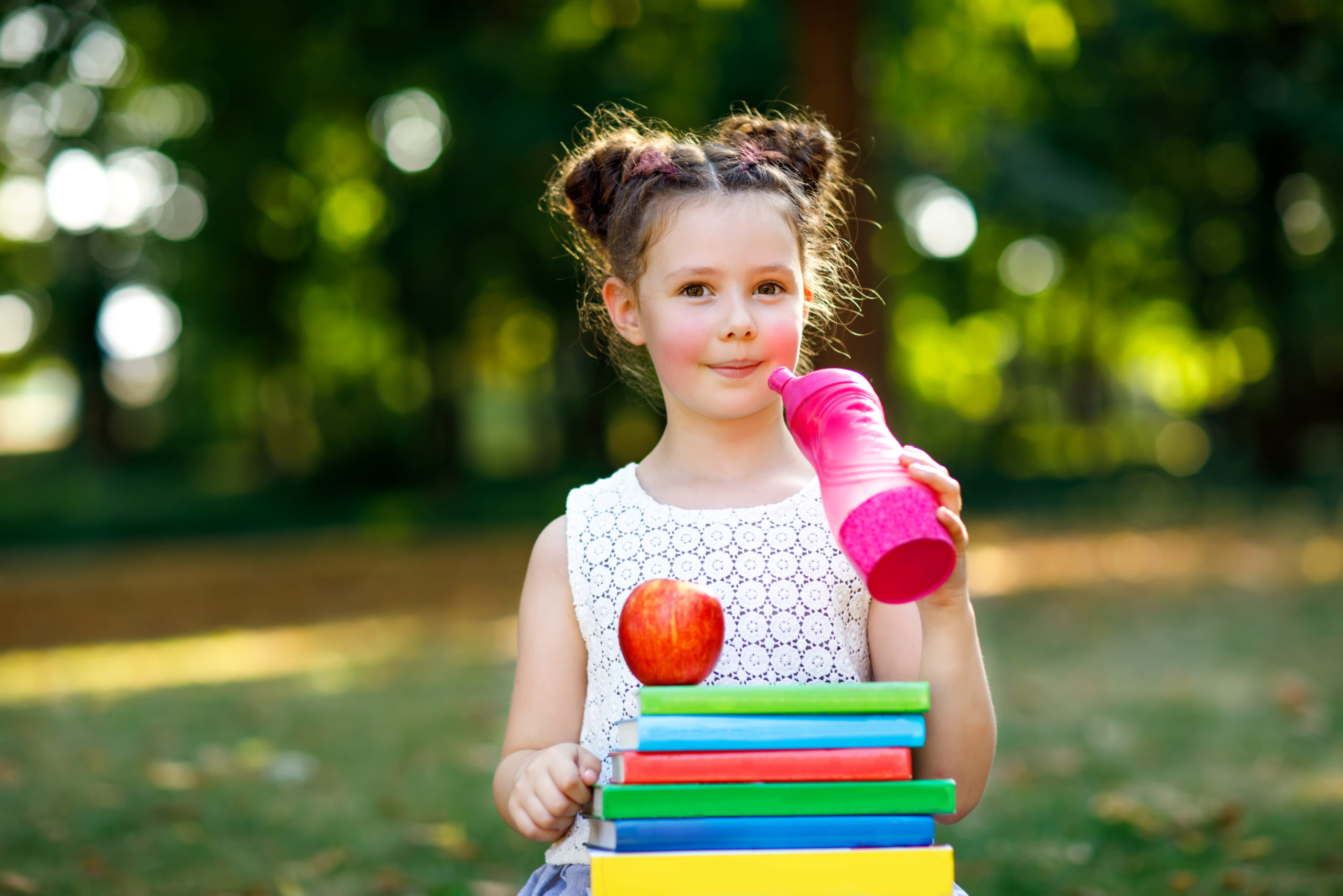 Mezzobusto di bambina in piedi all'aperto con una pila di libri davanti sulla quale è appoggiata una mela, mentre tiene in mano e avvicina alla bocca una borraccia