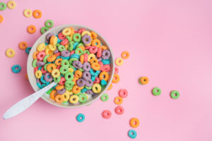 Cereali da colazione colorati in una tazza con cucchiaio, appoggiata su una superficie rosa; concept: alimenti ultra processati