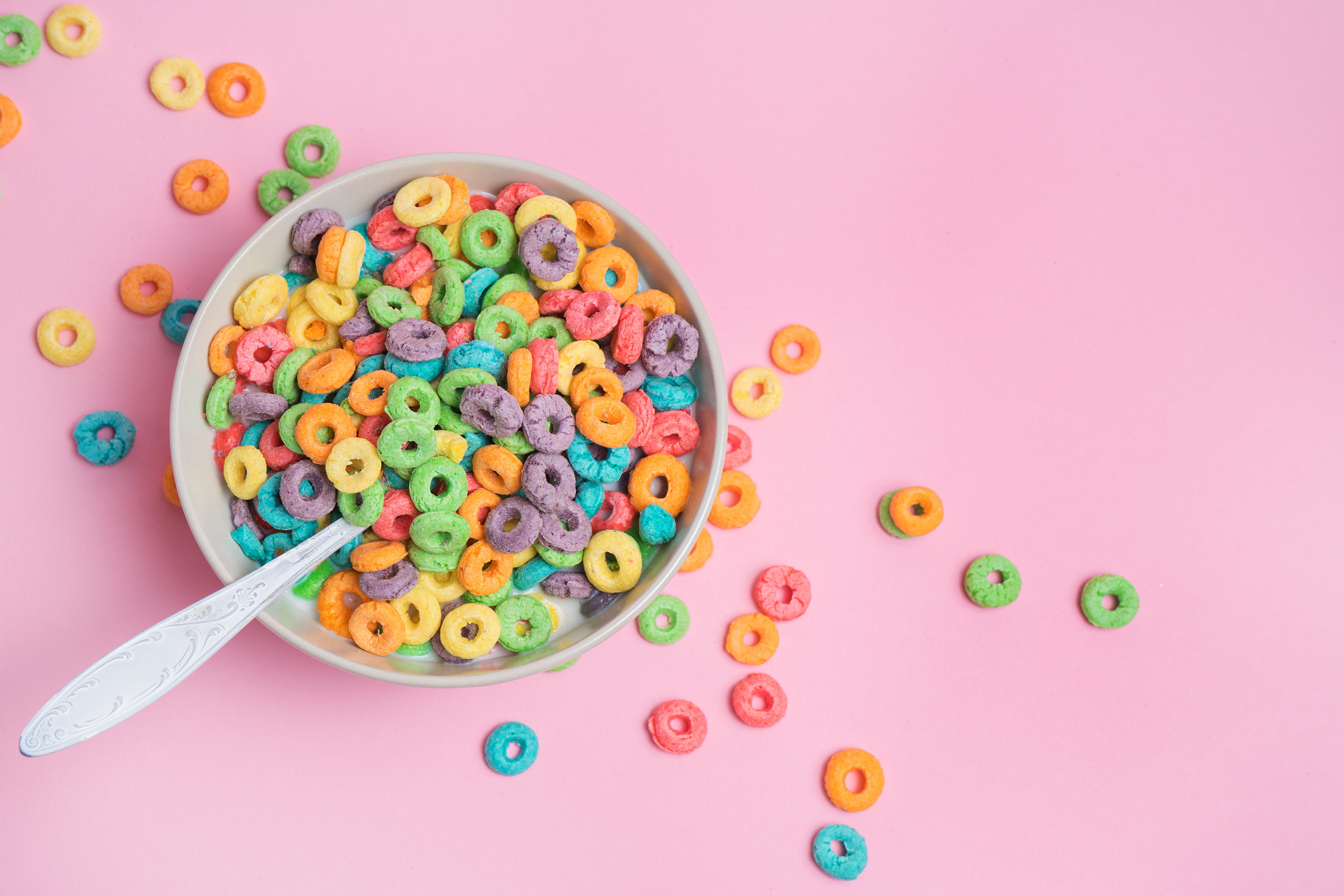 Cereali da colazione colorati in una tazza con cucchiaio, appoggiata su una superficie rosa; concept: alimenti ultra processati