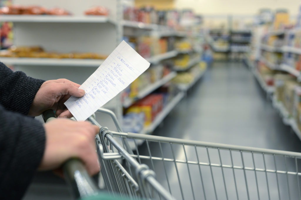 Persona spinge carrello tra le corsie di un supermercato con la lista della spesa in mano