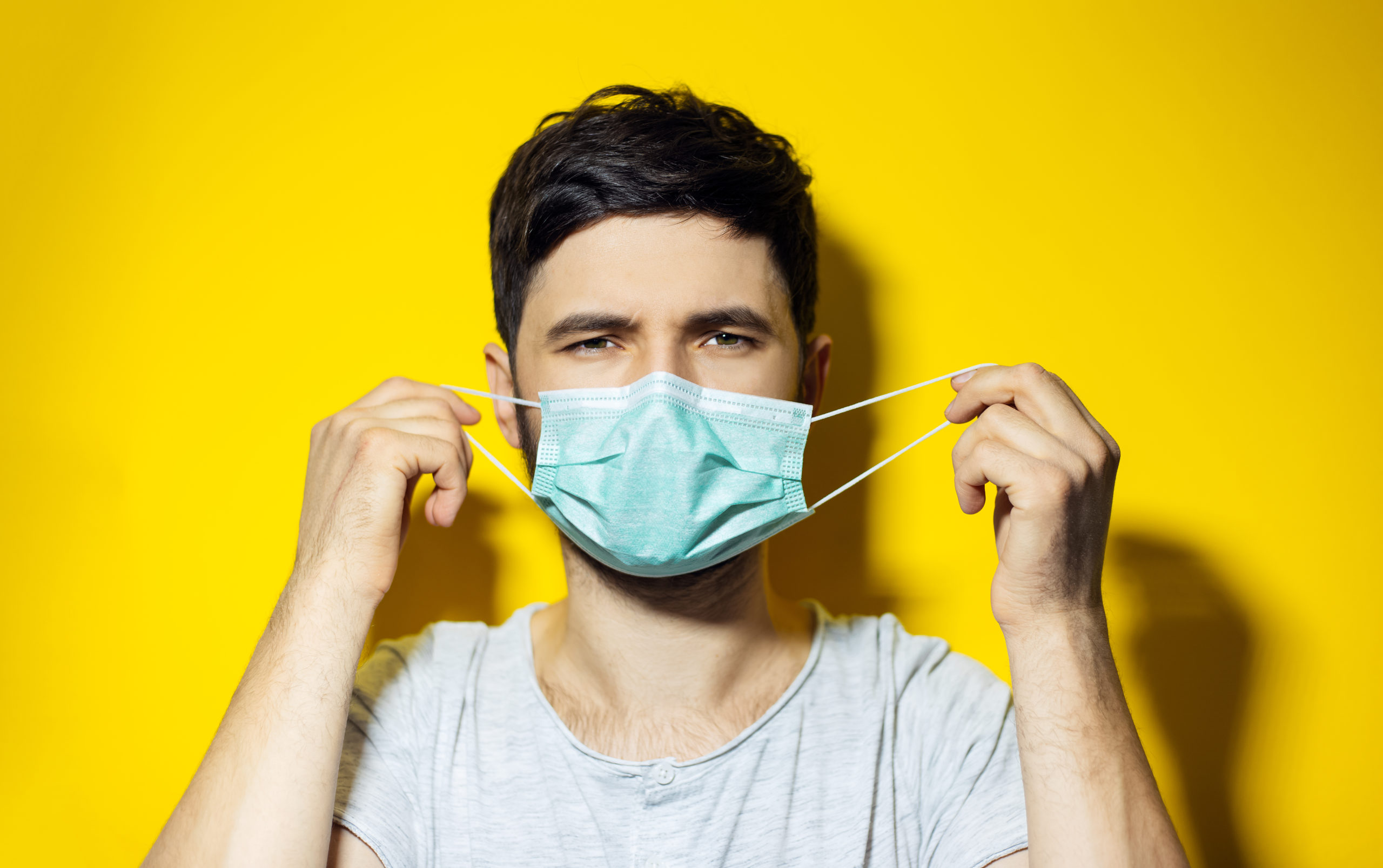 Studio portrait of young ill man, trying to put on, medical flu mask on yellow background mascherine