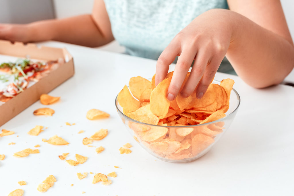 Breaking Diet. Chubby girl sitting at kitchen table with pizza eating chips close-up