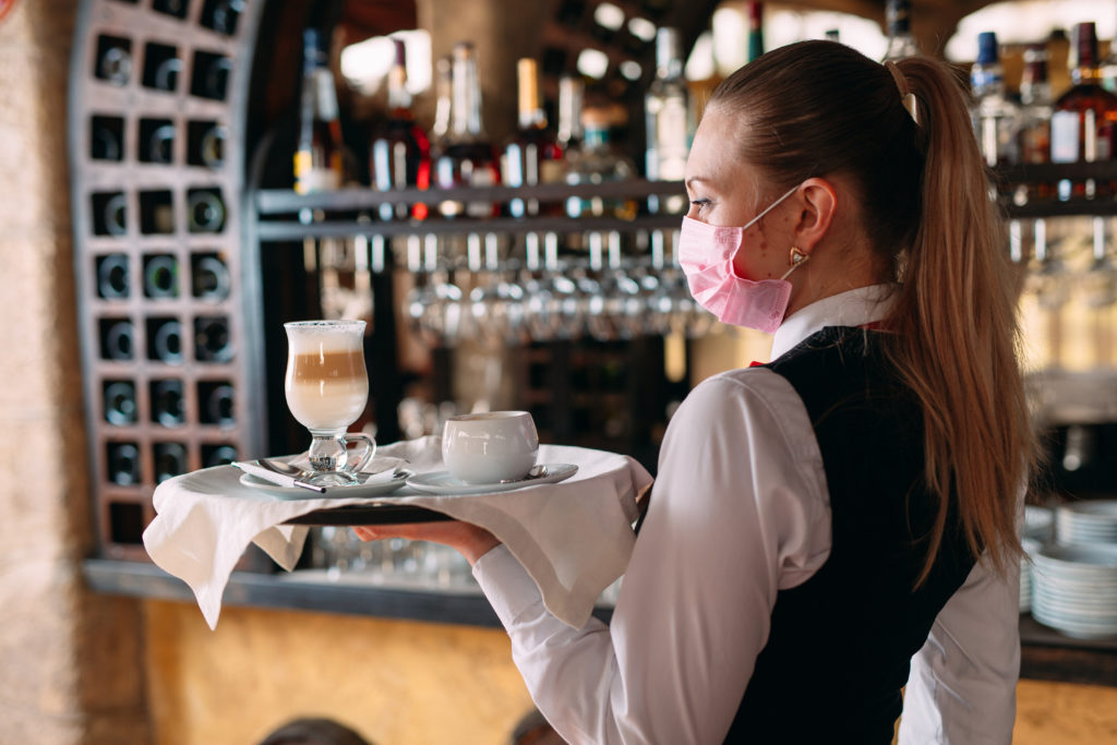A female Waiter of European appearance in a medical mask serves Latte coffee.