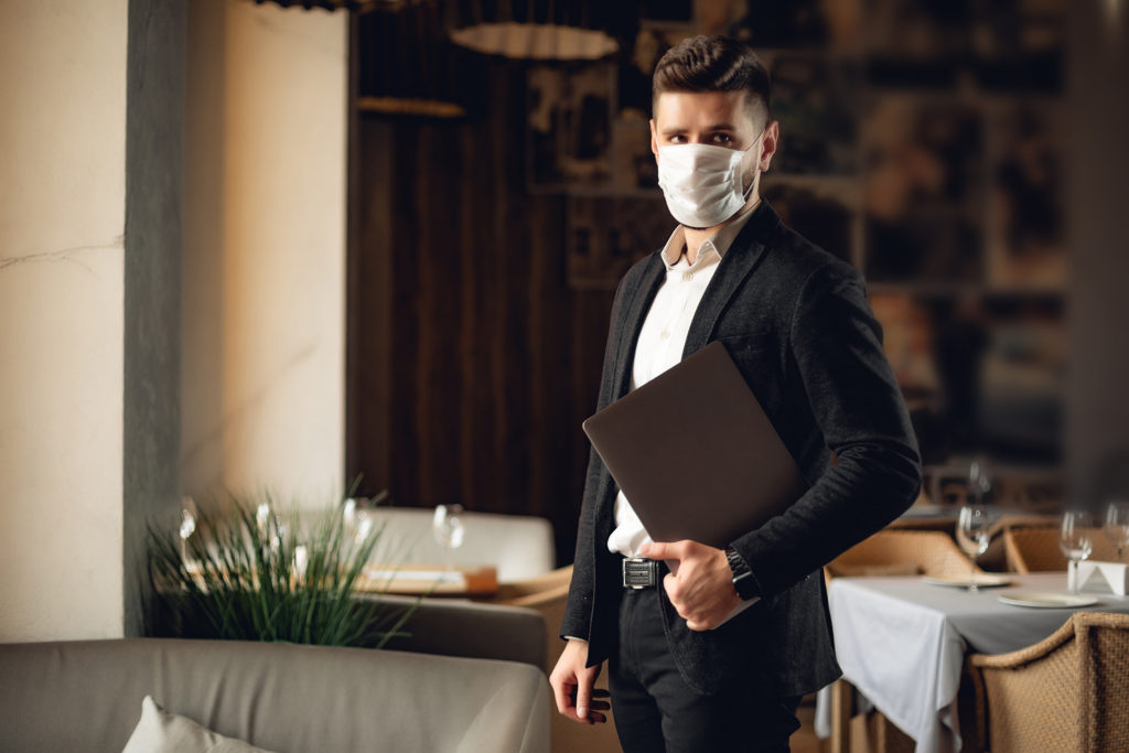 Handsome adult bearded man indoors in cafe. Lifestyle concept photo with copy space. Picture with gray laptop and protective mask on the face casual eating