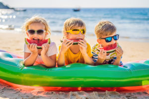 Bambini con gli occhiali da sole mangiano fette di anguria appoggiati su un gonfiabile in spiaggia, in riva al mare
