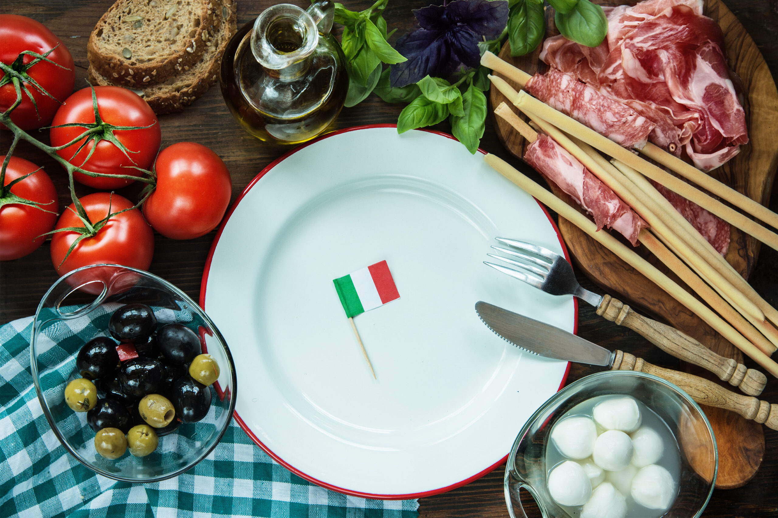 Piatto con bandiera italiana, circondato da prodotti tipici: ciotola di olive, pomodori, pane, olio extravergine di oliva, basilico, grissini, salame e ciotola di mozzarelline; concept: origine, cibo italiano, made in Italy