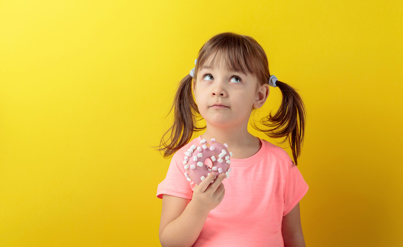 Bambina pensierosa con una ciambella glassata o donut in mano; concept: bambini, dolci, merendine