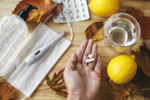 Una mano regge capsule di farmaci o integratori su un tavolo con bicchiere d'acqua, limoni, blister di pastiglie, termometro, mascherina; concept: influenza, raffreddore, covid-19, coronavirus, sistema immunitario, vitamine