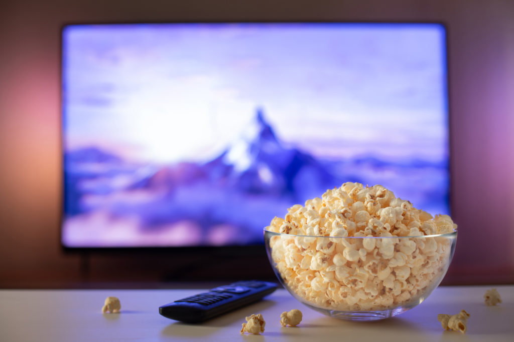 A glass bowl of popcorn and remote control in the background the TV works. Evening cozy watching a movie or TV series at home