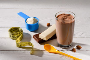 Prodotti protein/proteici: bicchiere con bevanda al cioccolato o caffè o nocciola, barrette, polvere in misurino blu, cucchiaino arancione e metro; concept: pasti sostitutivi
