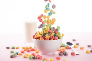 Cereali da colazione per bambini che cadono in una tazza, sullo sfondo un bicchiere di latte; concept: zucchero, coloranti