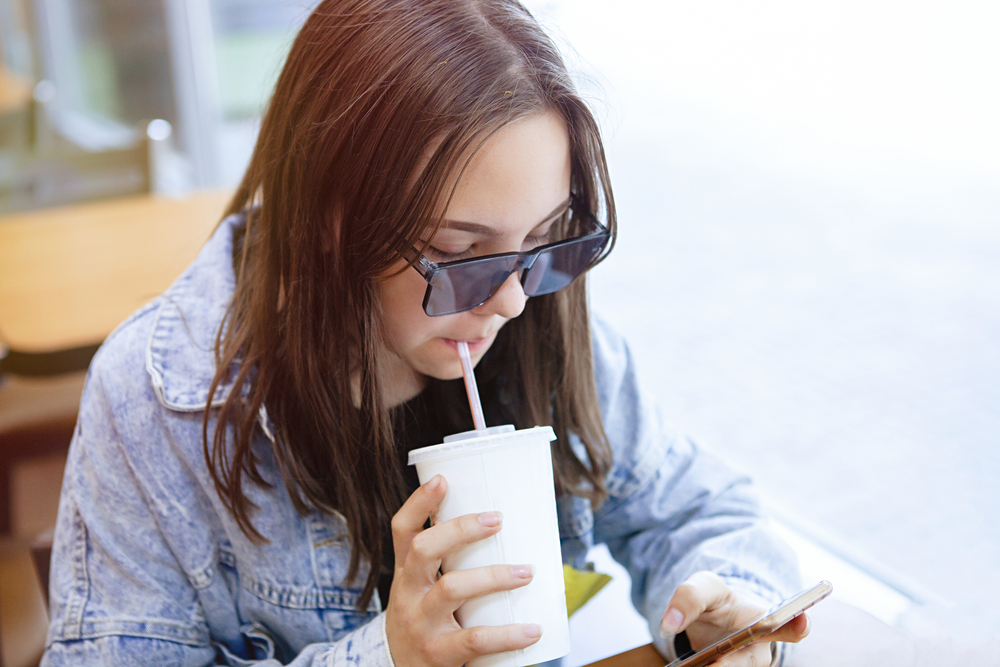 Ragazza adolescente beve una bevanda zuccherata con una cannuccia mentre guarda lo smartphone