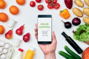 Una mano tiene uno smartphone con il logo di un carrello e la scritta online shopping, tutto intorno frutta, verdura e altri prodotti non alimentari; concept: spesa online, prezzi