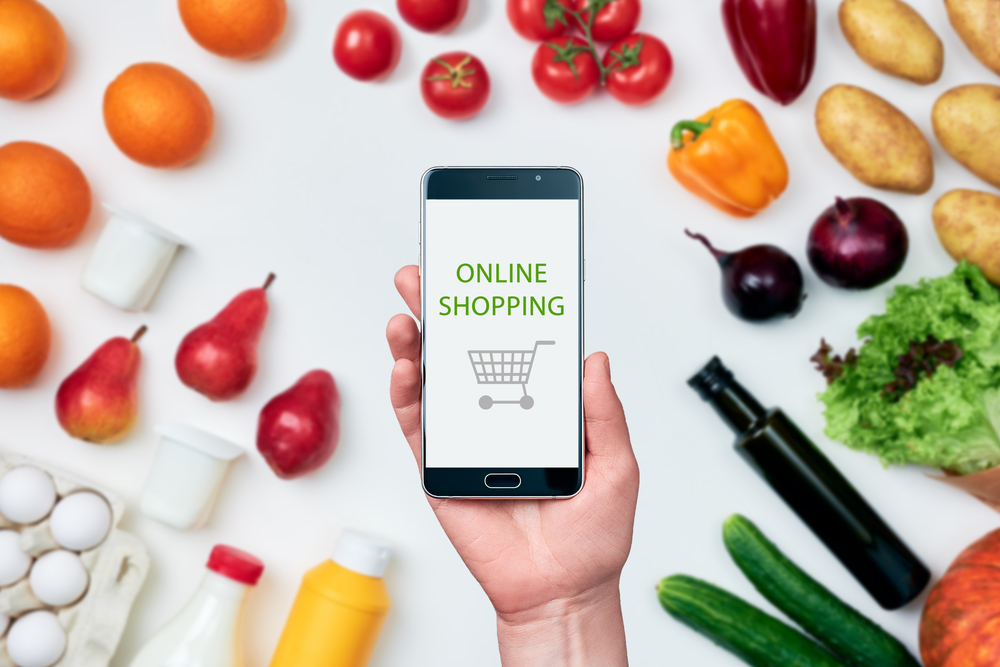 Una mano tiene uno smartphone con il logo di un carrello e la scritta online shopping, tutto intorno frutta, verdura e altri prodotti non alimentari; concept: spesa online, prezzi
