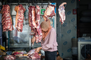 pezzi di carne appesi sopra il banco di un mercato, sotto una venditrice seziona altra carne
