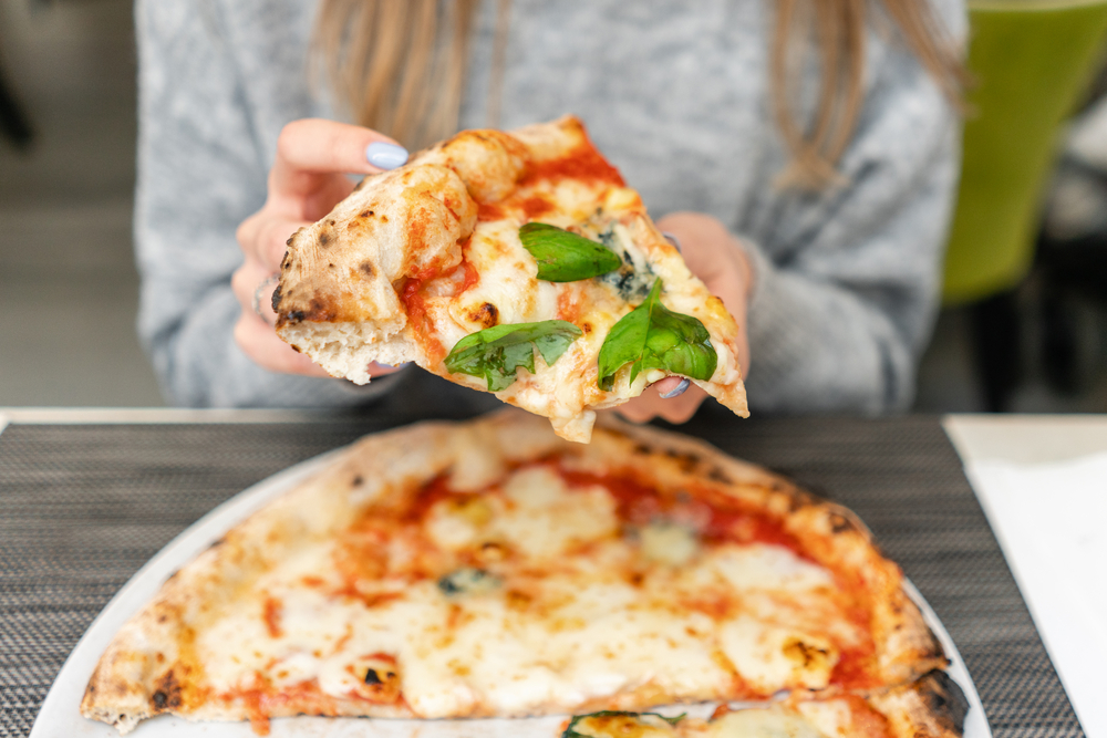 Una donna tiene in mano una fetta di pizza margherita con basilico, sopra un piatto di pizza margherita; concept: pizza napoletana