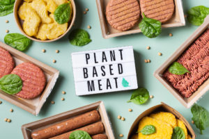 Assortimento di sostituti vegetali della carne (burger, salsicce, trita, nugget e cotolette) attorno a una lavagnetta luminosa con la scritta “plant based meat”; concept: prodotti vegetali, prodotti vegani