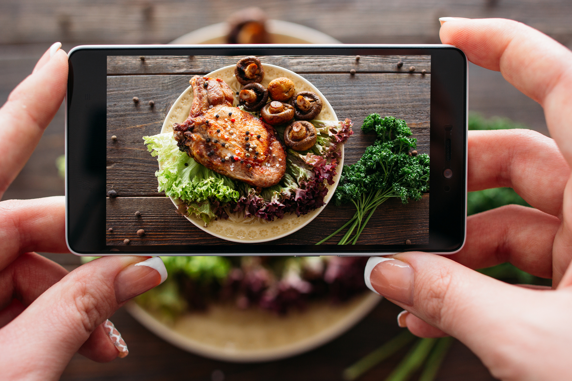 Smartphone tenuto da mani femminili inquadra piatto di carne di maiale con insalata e funghi, accanto al piatto mazzo di prezzemolo o coriandolo; concept social media