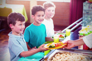 Bambini in fila al bancone della mensa scolastica mentre un'operatrice passa un piatto con carote, broccoli e pasta al primo della fila; concept: Milano Ristorazione