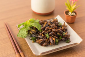 Grilli o cavallette fritti in un piattino con menta ed erba cipollina, accanto a bacchette, tazza e vaso con piantina; concept: insetti commestibili