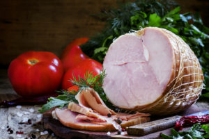 Prosciutto cotto in trancio e fette su un tagliere di legno con pomodori ed erbe sullo sfondo