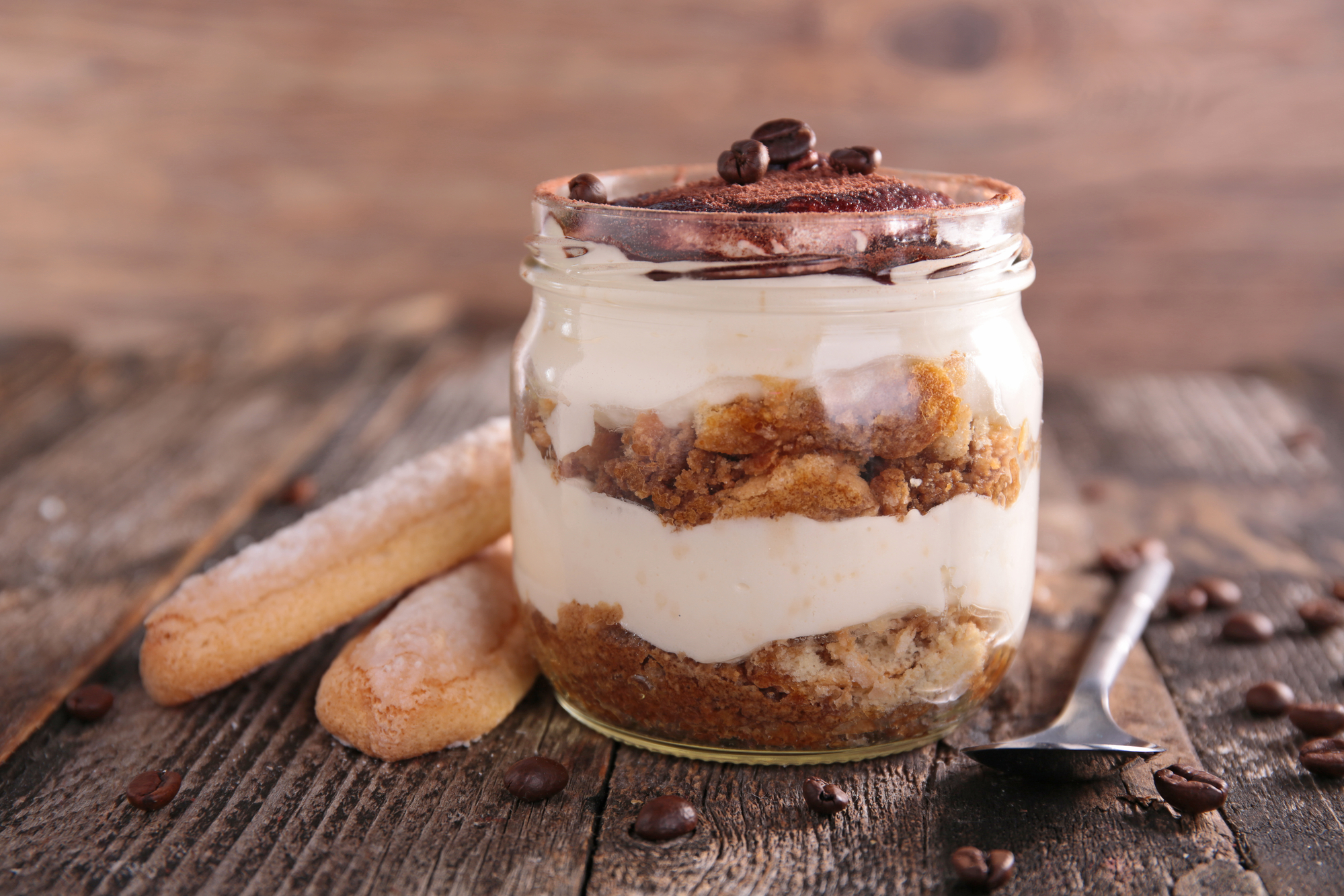 Tiramisù in barattolo di vetro su un tavolo di legno accanto a savoiardi e cucchiaino, con grani di caffè sparsi