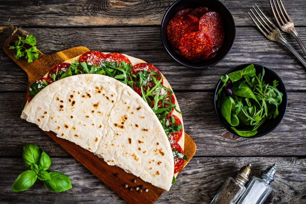 Piadina con salame e rucola su tagliere di legno, accanto a ciotole con salame e rucola, saliera e pepiera