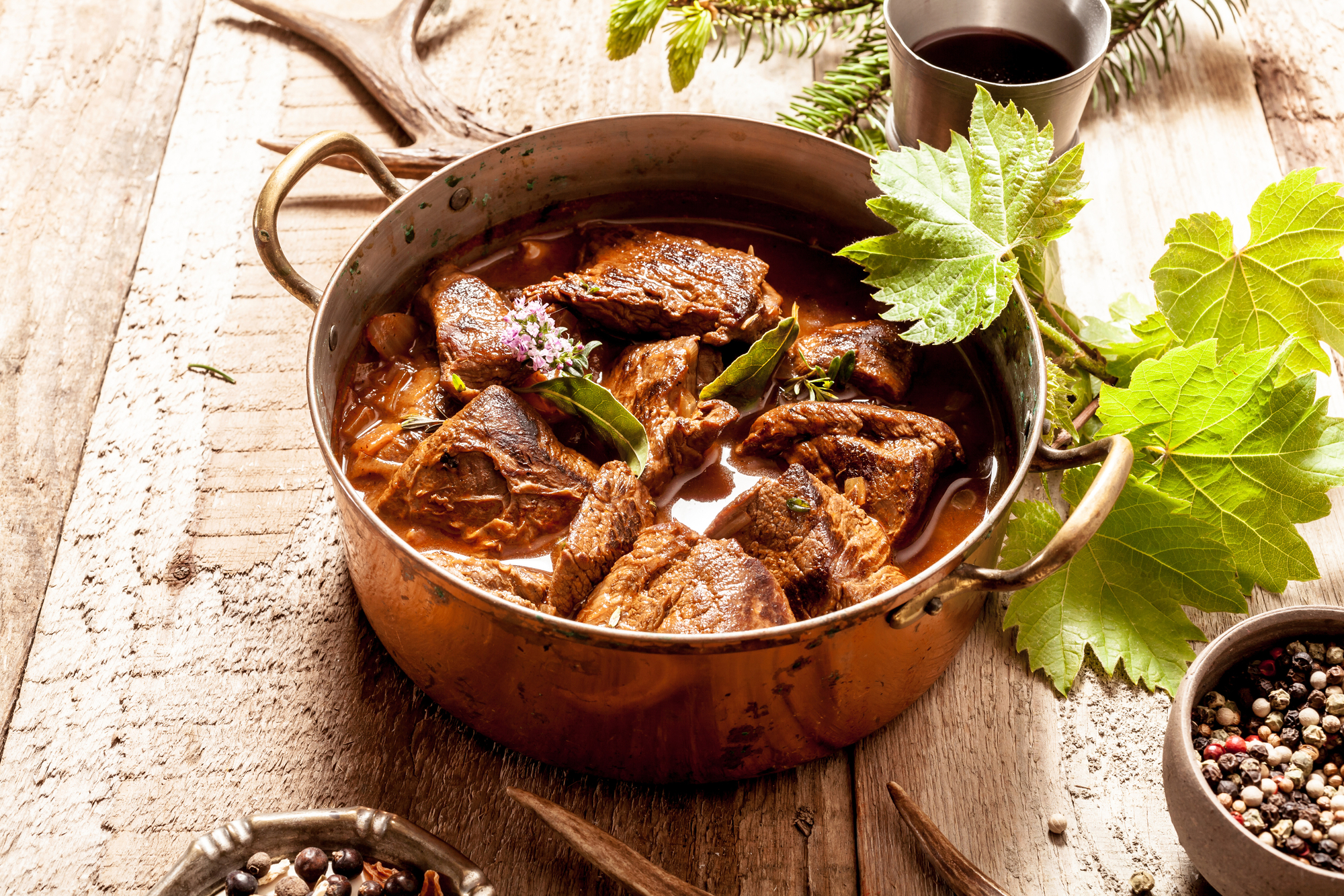 Gulasch di cervo stufato in pentola di rame con ciotole di condimento su una superficie di legno circondata da corna e foglie di cervo cinghiale carne proteine cucina piatto preparazione cucinare