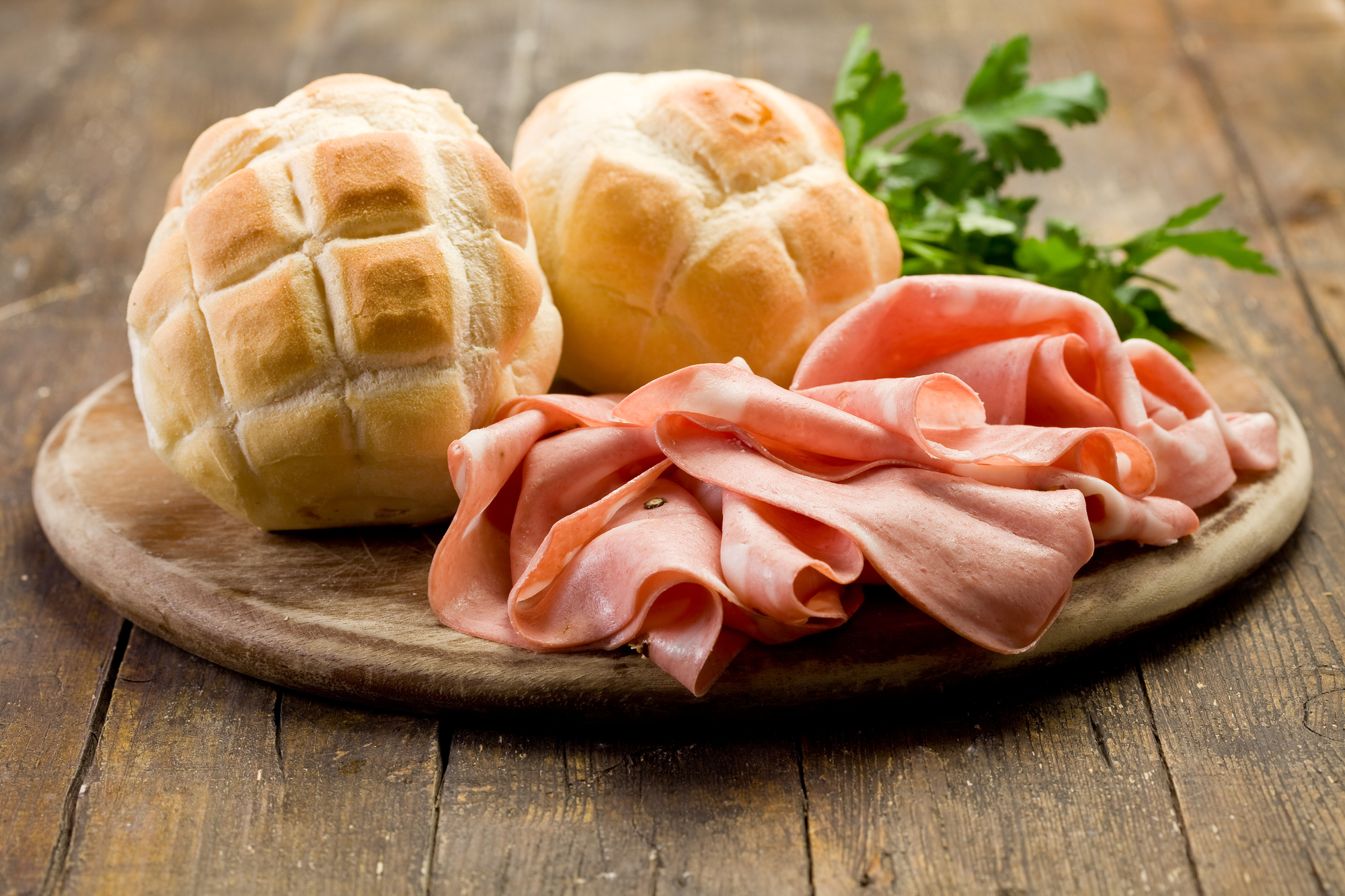 Fette di mortadella, due pagnotte e un ciuffo di prezzemolo su un tagliere o un piatto di legno