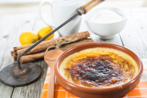 Crema catalana in una ciotola di coccio, con cucchiaio di legno, cannella, limone, latte, zucchero