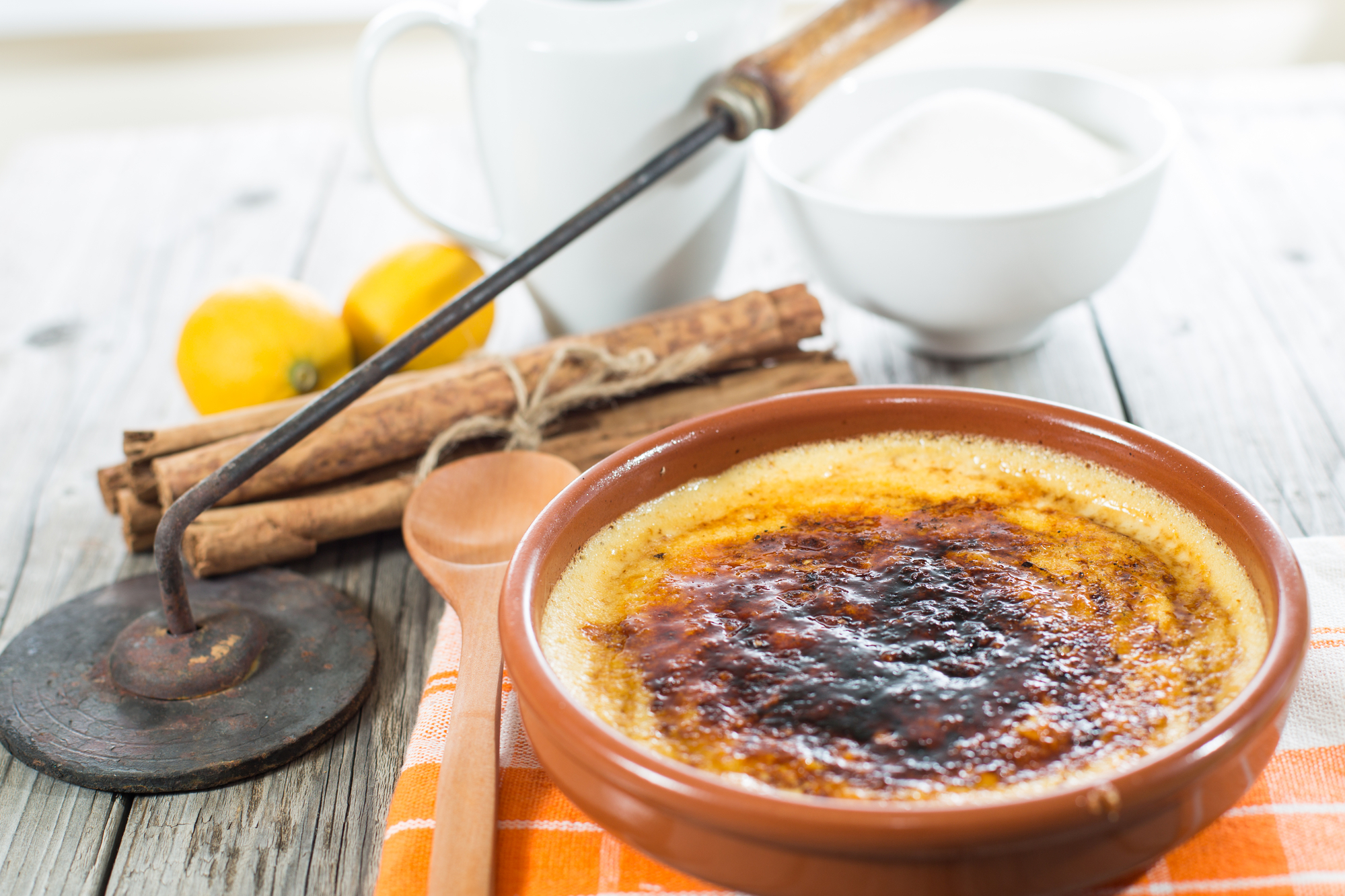 Crema catalana in una ciotola di coccio, con cucchiaio di legno, cannella, limone, latte, zucchero