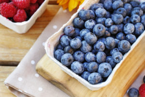 Mirtilli in una scatola di carta su un tagliere, sullo sfondo lamponi in confezione di carta