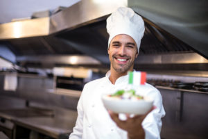 Chef o cuoco nella cucina di un ristorante italiano tiene in mano piatto di pasta con bandiera italiana