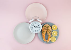 Due piatti vuoti e uno con salmone e fette di limone, con al centro una sveglia; concept: digiuno intermittente