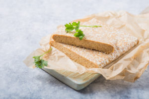 Tempeh con erbe su foglio di carta da forno appoggiato su un tagliere di legno chiaro; concept: sostituti vegetali, vegan
