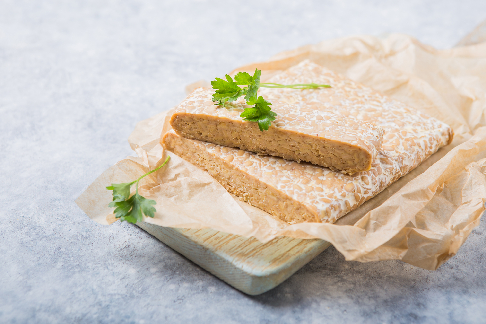 Tempeh con erbe su foglio di carta da forno appoggiato su un tagliere di legno chiaro; concept: sostituti vegetali, vegan