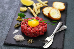 Tartare di bovino con tuorlo d'uovo su un vassoio di ardesia insieme a cipolla, sale, capperi, erbe, crostini e limone