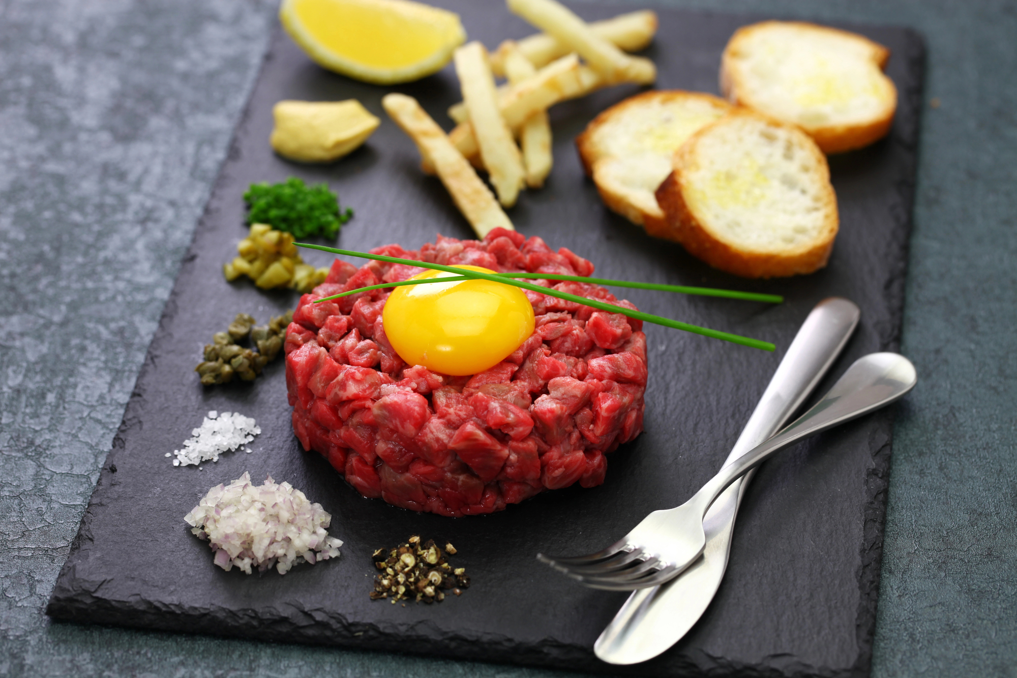 Tartare di bovino con tuorlo d'uovo su un vassoio di ardesia insieme a cipolla, sale, capperi, erbe, crostini e limone