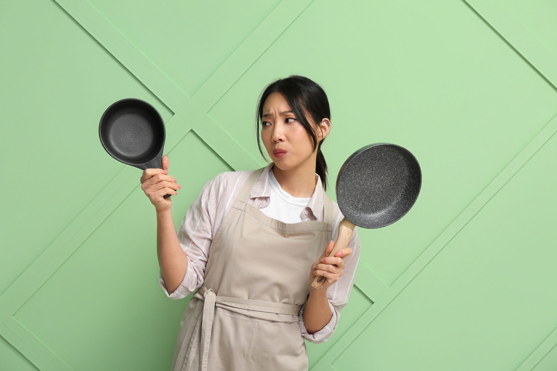 Donna asiatica con grembiule da cucina e sguardo dubbioso tiene in mano una padella antiaderente e una padellina di ghisa; concept: padelle, cucina