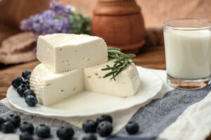 Forma di formaggio fresco di capra, primo sale o primosale tagliata su un piattino bianco con rosmarino e mirtilli, accanto a un bicchiere di latte, sotto il piatto uno strofinaccio con mirtilli sparsi
