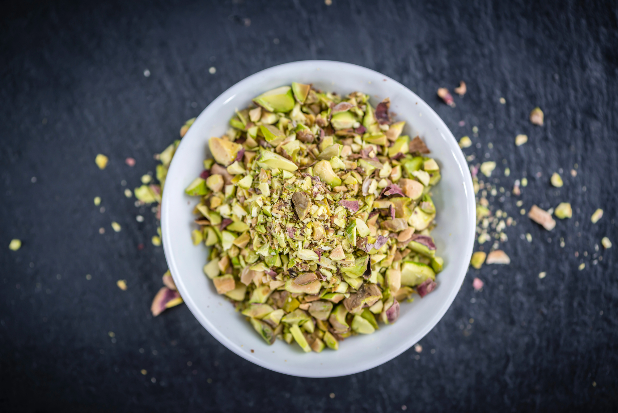 Granella di pistacchi in una ciotolina bianca su una superficie nera con altra granella sparsa; concept: frutta secca