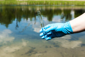 Una persona con guanto blu sulla mano tiene una provetta di acqua prelevata da un fiume o un laghetto; concept: inquinamento delle acque, PFAS, analisi