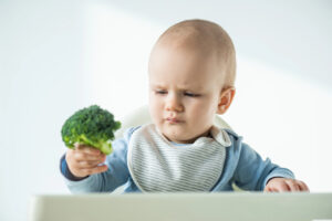 Un bambino piccolo con bavaglino seduto su un seggiolone osserva con sguardo dubbioso la cimetta di un broccolo; concept: svezzamento, frutta e verdura
